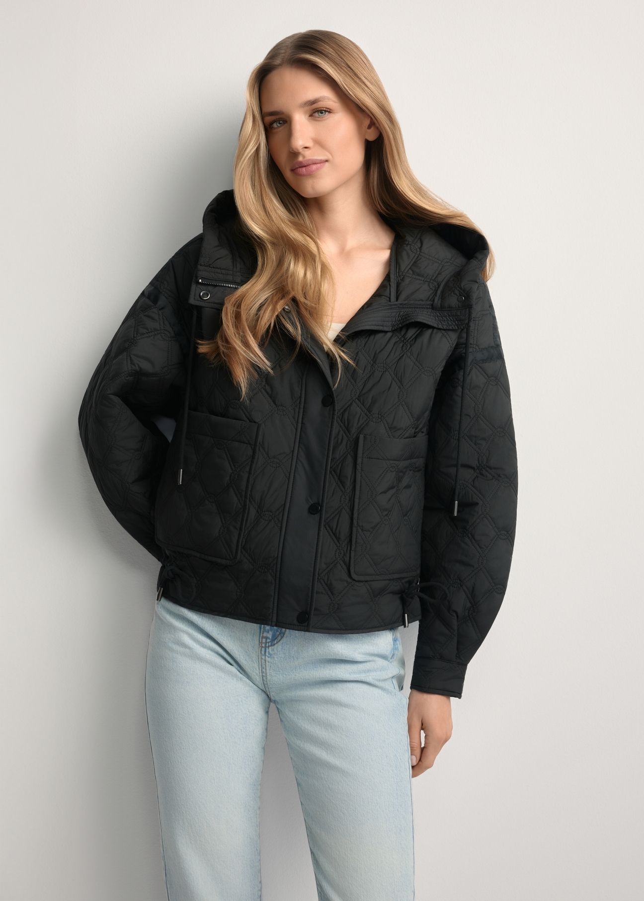 Czarna krótka pikowana kurtka oversize KURDT-0660-99(W26) - widok produktu z przodu, na modelce, na jasnym tle. Widoczna sylwetka do kolan. 
